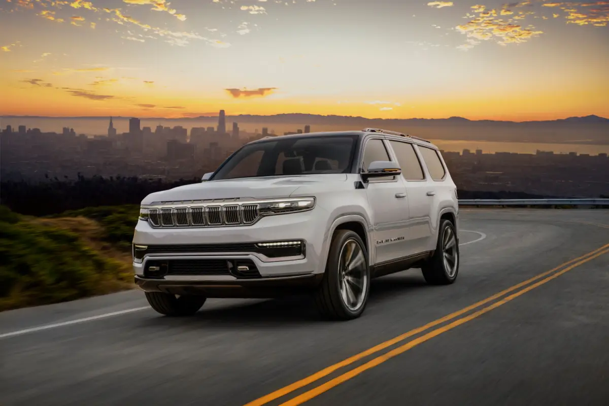 White Jeep Grand Wagoneer full-size luxury SUV driving on a winding road overlooking a city skyline at sunset