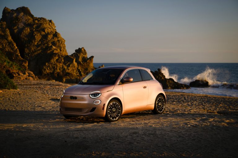 2024 Rose gold Fiat 500e electric car parked on a beach with ocean waves in the background