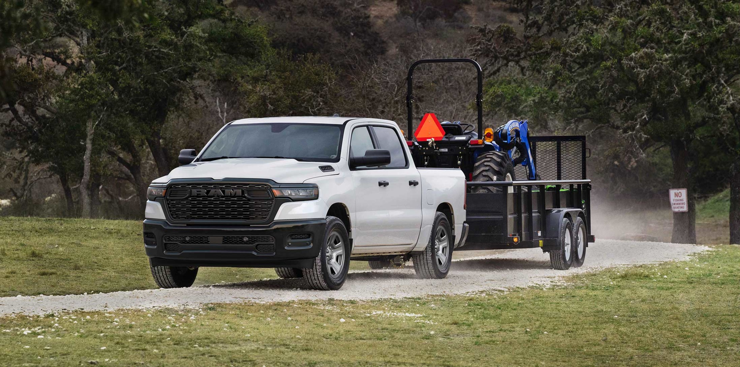2026White 2025 Ram 1500 pickup truck towing a New Holland tractor on a utility trailer down a gravel path