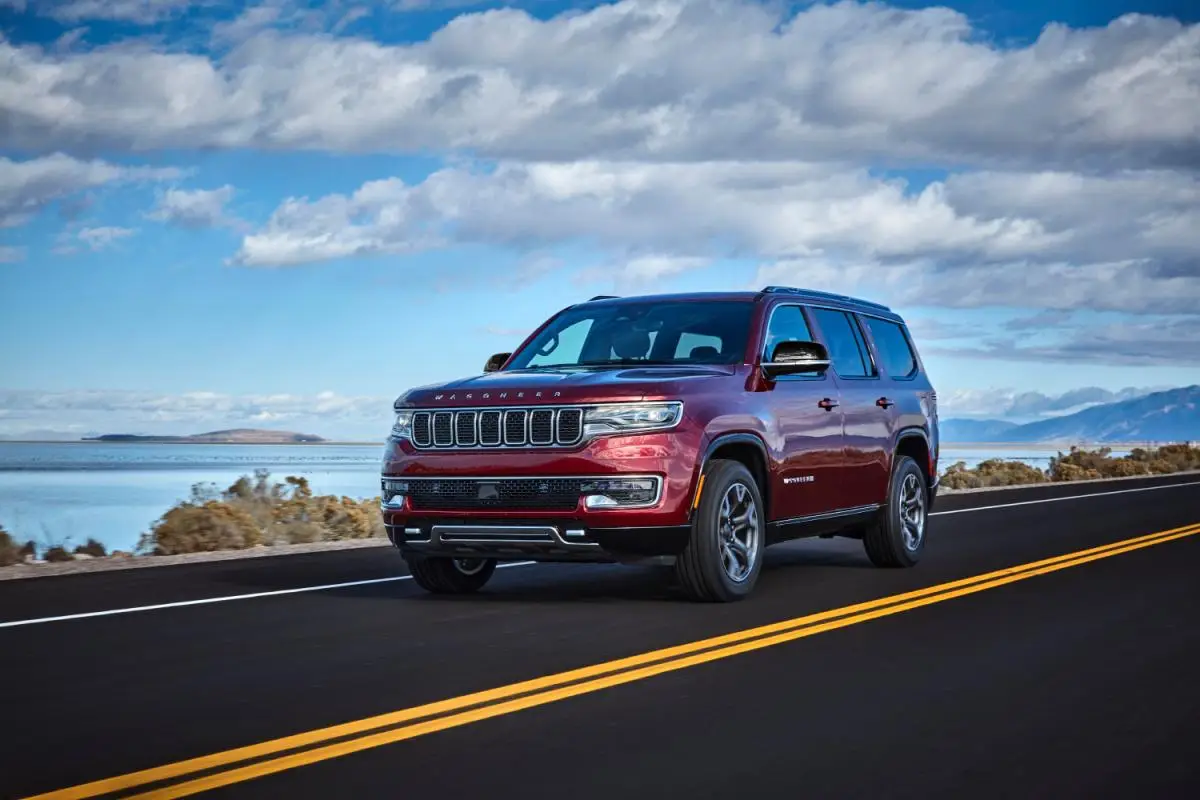 2023 Red Jeep Wagoneer full-size SUV driving on a coastal road beside a lake, front three-quarter view