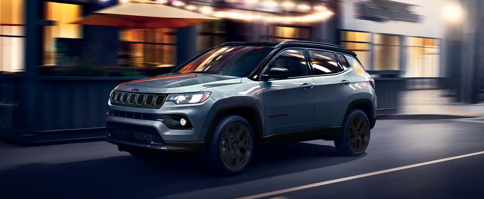 Gray 2025 Jeep Compass SUV driving through a city street at dusk with illuminated storefronts